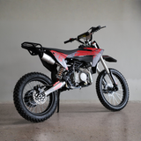 A red and black off-road dirt bike with a silver exhaust and knobby tires, photographed on a concrete floor against a plain gray wall.