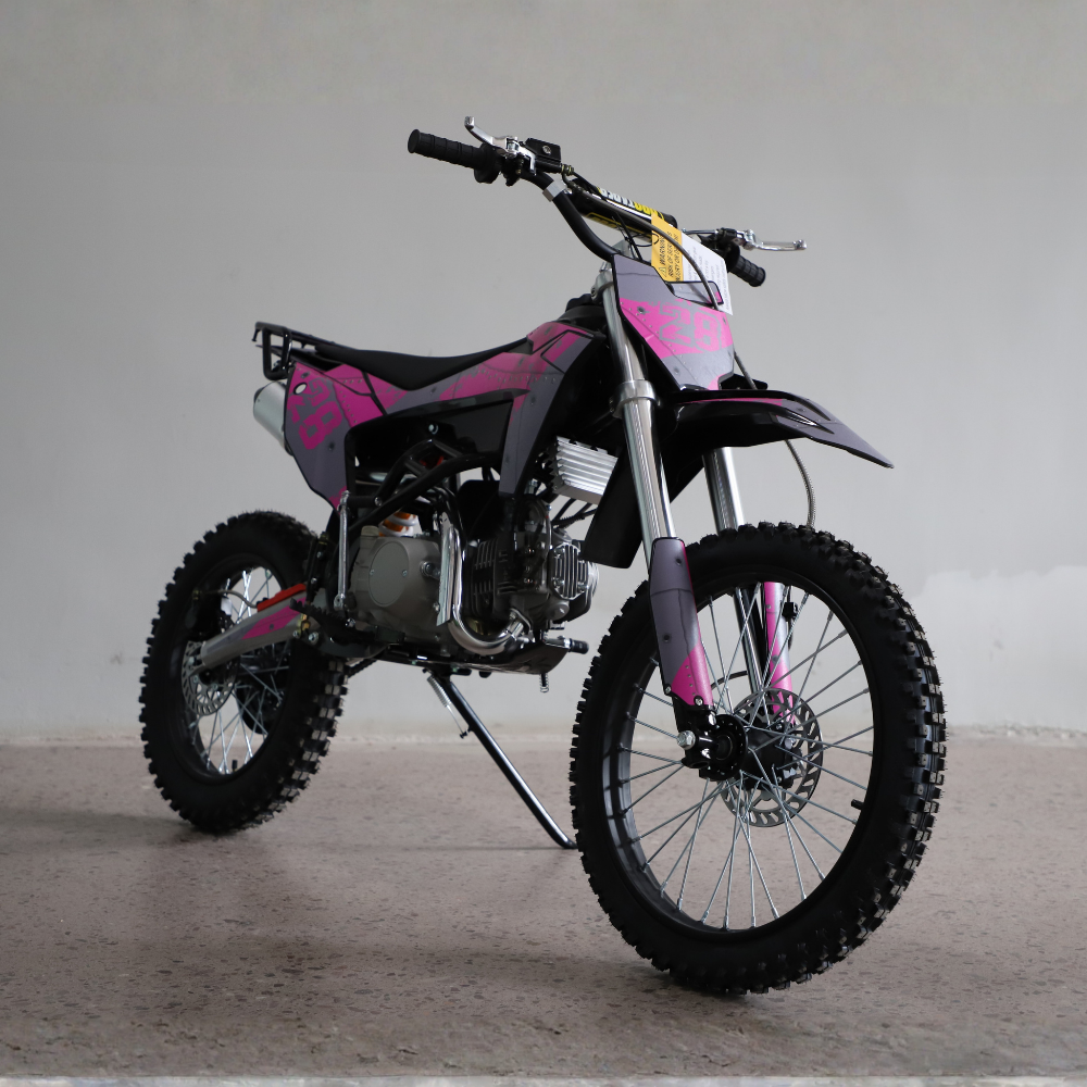 A pink and black mini dirt bike with knobby tires, photographed on a concrete floor against a plain gray wall.