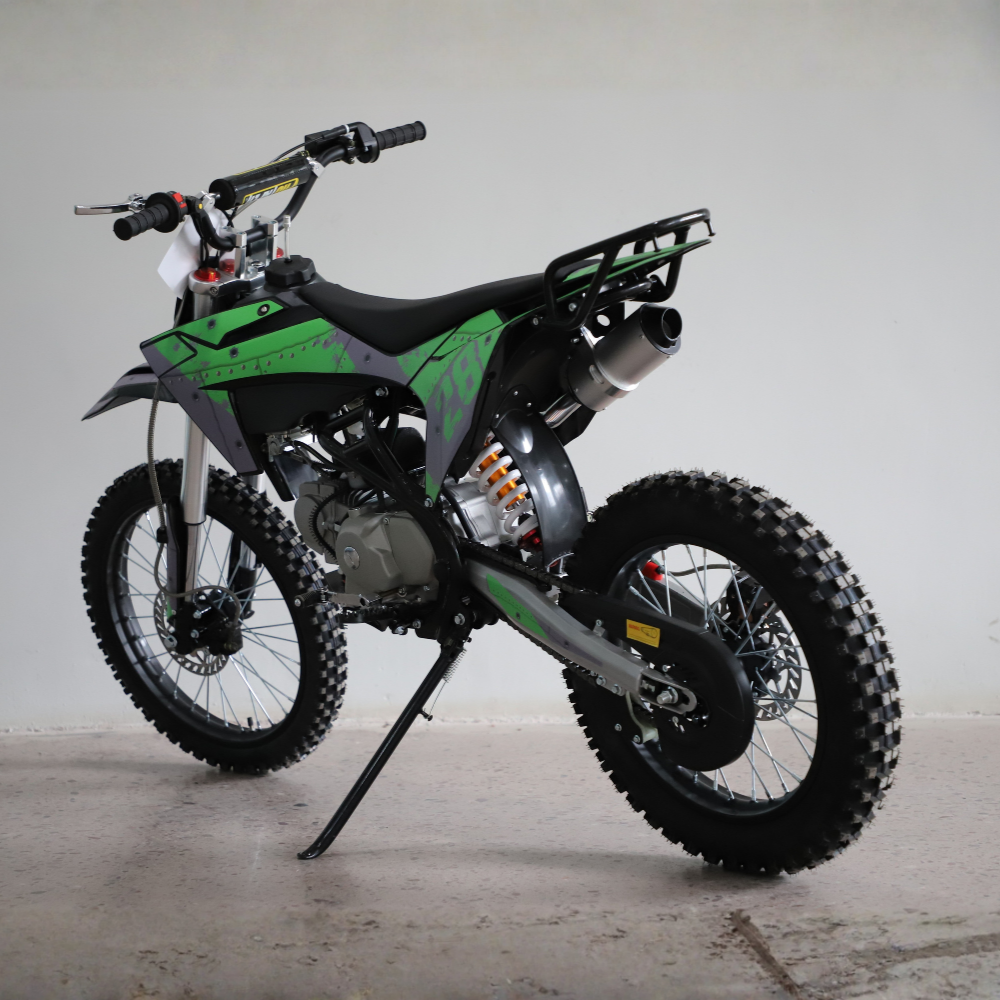 A green and black dirt bike with knobby off-road tires, a black seat, and a silver exhaust pipe, shown on a kickstand against a plain light gray wall.