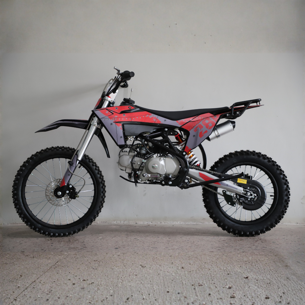 A red and black dirt bike with a silver engine and black knobby tires, photographed from the side against a plain concrete wall and floor.