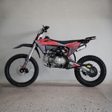 A red and black dirt bike with a silver engine and black knobby tires, photographed from the side against a plain concrete wall and floor.