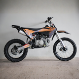 A 125cc dirt bike with orange and black graphics, silver forks, and knobby tires, photographed from a side profile against a plain concrete wall.