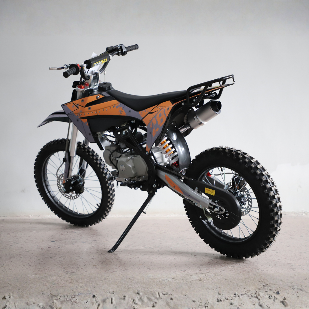 A small off-road dirt bike with orange and gray graphics, black seat, knobby tires, and a black kickstand, photographed indoors against a plain light gray wall and concrete floor.
