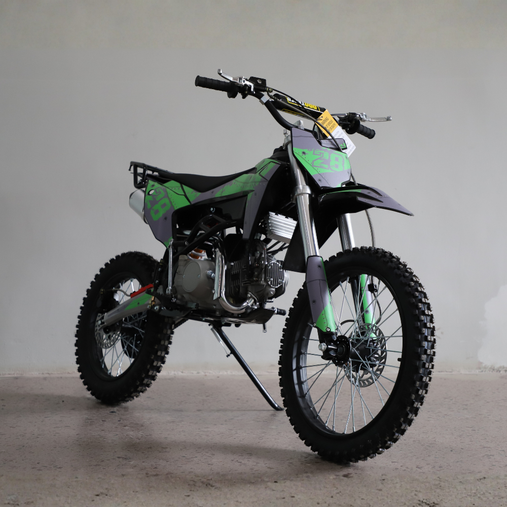 A small off-road dirt bike with knobby tires, a black seat, and green and black graphics, photographed indoors against a plain light gray wall and concrete floor.