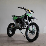 A small off-road dirt bike with knobby tires, a black seat, and green and black graphics, photographed indoors against a plain light gray wall and concrete floor.
