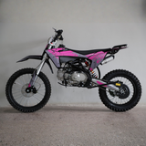 A pink and black mini dirt bike with knobby tires, photographed from the side against a plain gray wall and concrete floor.