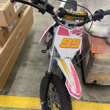 Pink and white Motoworks 90cc petrol-powered kids' dirt bike with number 99, standing in a warehouse next to cardboard boxes.