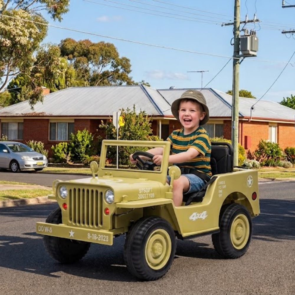 A young boy wearing a safari hat drives a small military-style toy jeep on a suburban street. The toy jeep is olive green with black tires and seats, and has decorative white and red markings.