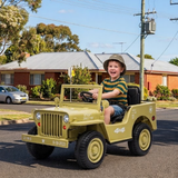 A young boy wearing a safari hat drives a small military-style toy jeep on a suburban street. The toy jeep is olive green with black tires and seats, and has decorative white and red markings.