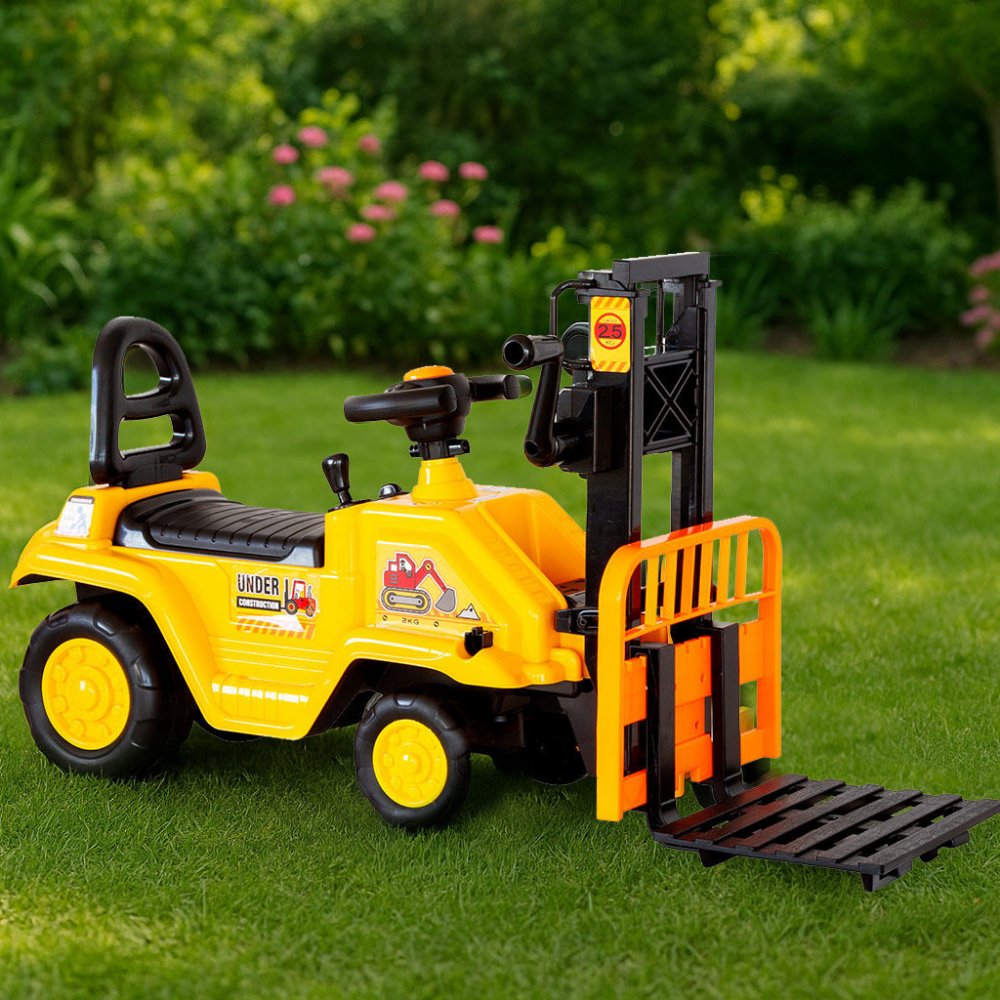 A yellow and black ride-on toy forklift with a front pallet, photographed outdoors on green grass with a blurred garden background.