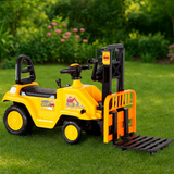 A yellow and black ride-on toy forklift with a front pallet, photographed outdoors on green grass with a blurred garden background.