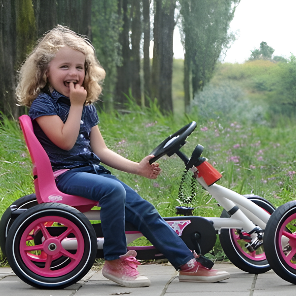 Child smiling and sitting in colourful pedal-powered go-karts, one pink and white, enjoying an outdoor activity on a paved path surrounded by greenery