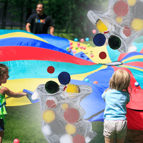 Children playing with a colourful parachute while fleece balls in various colours are being tossed into the air, with some fleece balls shown in a mesh bag in the foreground