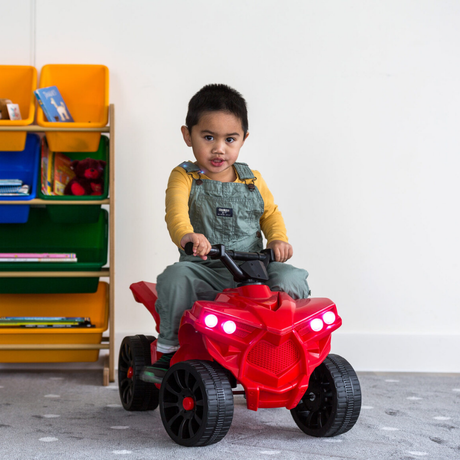 Child sitting on a red toy ATV in a room with colorful shelves.