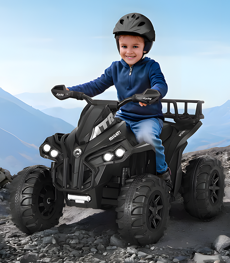 A young child wearing a helmet is smiling while riding a black four-wheeler toy ATV. The ATV is designed for rough terrain, with large rugged tyres and headlights. The background shows a mountainous outdoor landscape, enhancing the adventurous atmosphere