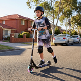 Child riding a scooter on a residential street with houses and cars in the background.