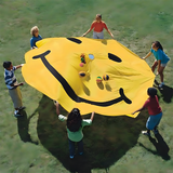 Children holding a yellow smiley face parachute while tossing small balls into the air during outdoor play