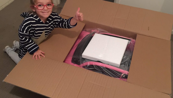 A young child wearing red glasses is smiling and giving a thumbs up while kneeling next to an open box containing a packaged item, possibly a toy or ride-on car