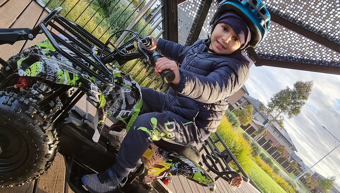 A young child in a blue helmet and winter jacket is sitting on a toy quad bike, ready to ride outdoors