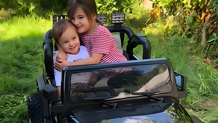 Two young children are seated in a black toy car outdoors, with the older child hugging the younger one while they enjoy a ride together