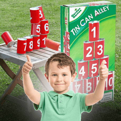 A young boy smiling and giving two thumbs up, standing in front of a Tin Can Alley game setup outdoors. The game consists of red numbered cans (1 to 10) stacked on a wooden table, with some cans knocked over, showcasing the fun of the game. The packaging of Tin Can Alley is also visible in the background, adding context to the product being promoted. The setting is a grassy outdoor area, suggesting a family-friendly activity perfect for the garden