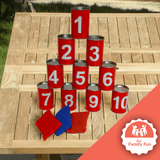 A set of red numbered cans (1 to 10) stacked in a pyramid shape on a wooden surface, with three coloured throwing bags (red and blue) placed in front. A badge in the bottom right corner reads "For Family Fun," indicating that the game is designed for enjoyable family activities. The setup suggests a fun, interactive outdoor game suitable for players of all ages