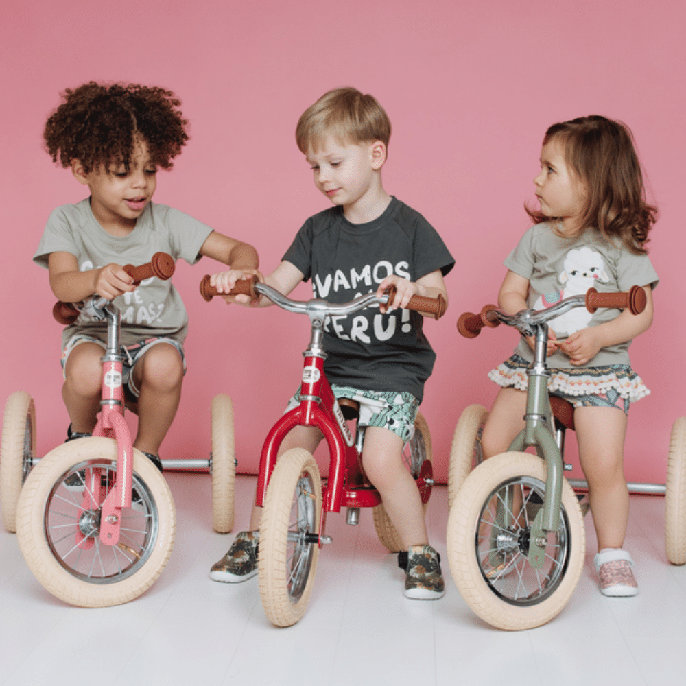 Three children riding balance bikes against a pink background
