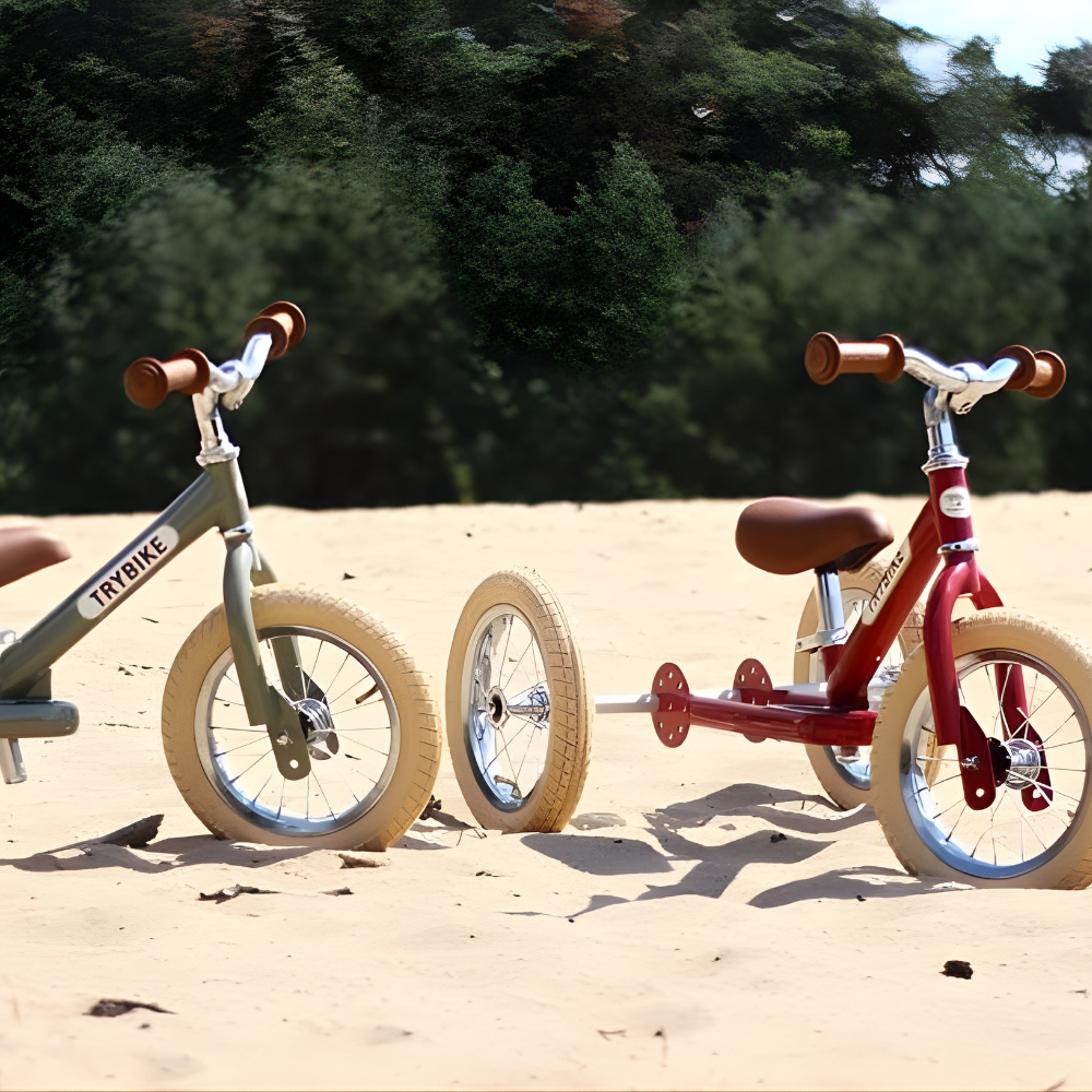 Two balance bikes, one green and one red, on a sandy surface with trees in the background.