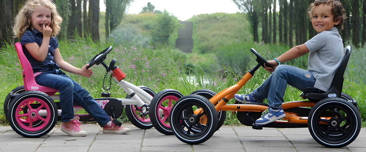 Two children smiling and sitting in colourful pedal-powered go-karts, one pink and white, the other orange and black, enjoying an outdoor activity on a paved path surrounded by greenery