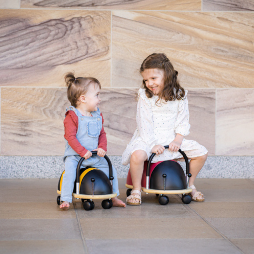 Two children sitting on small, round, black and yellow vehicles against a stone wall.