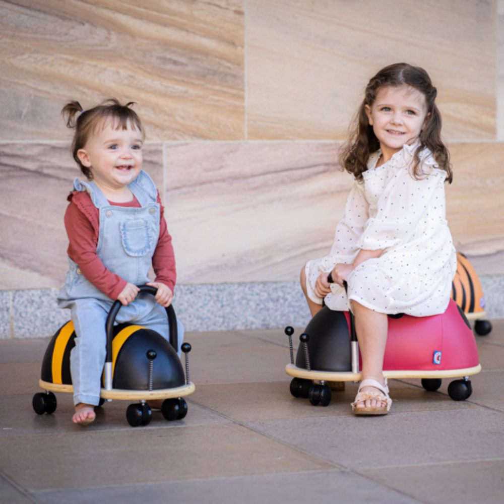 Two children sitting on colorful ride-on toys against a stone wall.