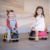 Two children sitting on colorful ride-on toys against a stone wall.