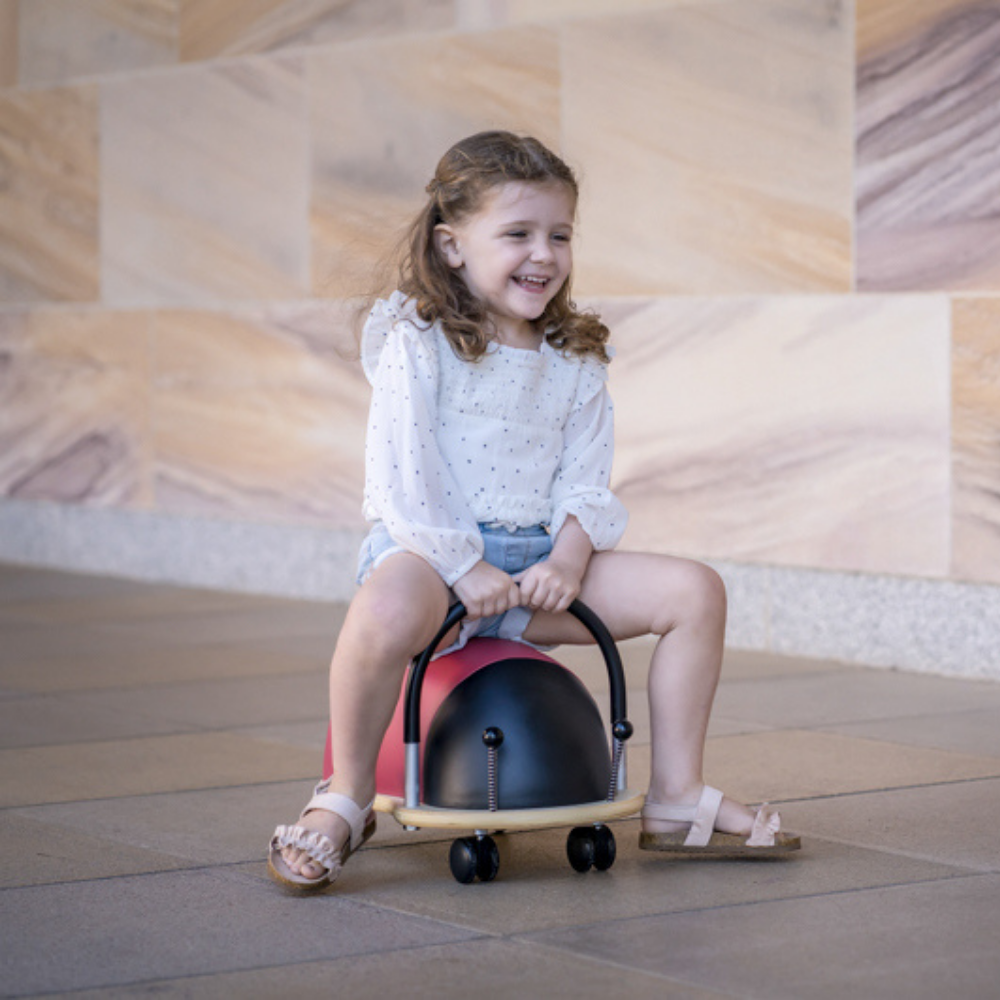 Young girl riding a colourful toy bug on a stone floor with a patterned wall in the background