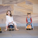 Two children sitting on small, round, black and orange ride-on toys against a stone wall.