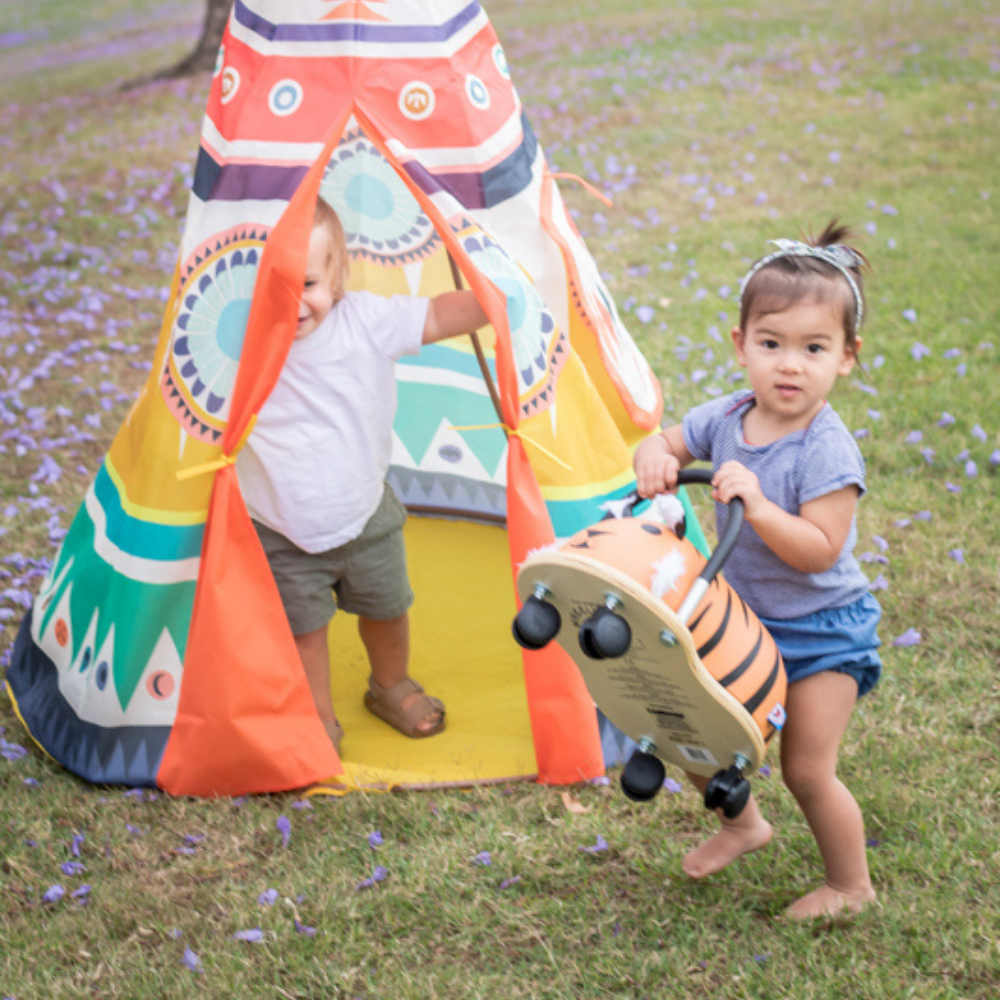 Two children playing with a toy car near a colorful teepee in a grassy area.
