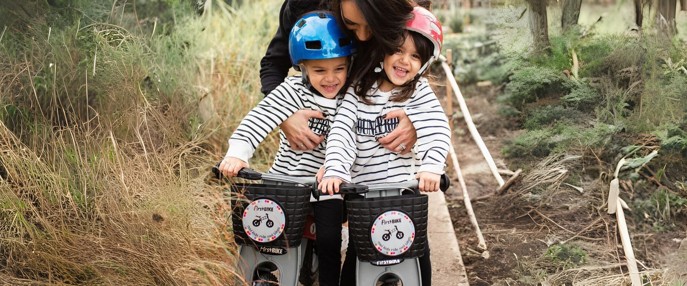 The image shows two young children, each wearing helmets (one blue and one pink), riding small balance bikes. They are wearing matching striped shirts and are smiling, clearly enjoying their outdoor activity. An adult, possibly a parent, is behind them, offering support and encouragement. The background is a nature trail surrounded by greenery, giving the impression of a family outing in a natural setting.