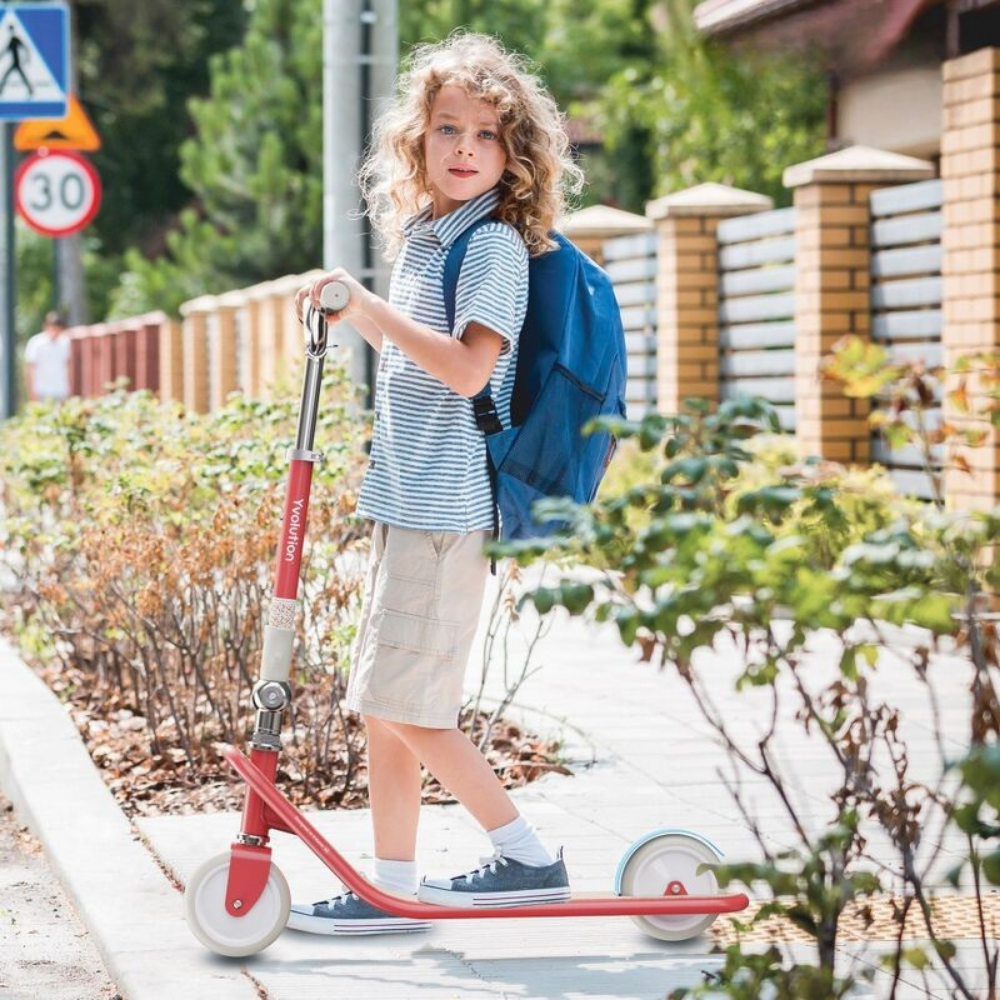Child with a red scooter and blue backpack standing on a sidewalk.