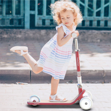 Child riding a red scooter on a sidewalk with a building in the background