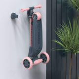 Children's scooter with pink and black design leaning against a wall next to a plant.