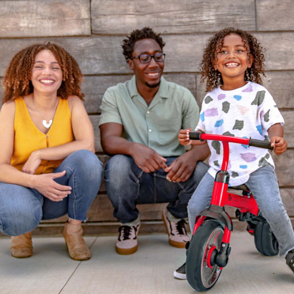 Family of three with a child on a red balance bike against a wooden wall.
