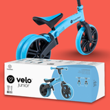 Blue balance bike with black wheels on a red background, next to its packaging box.
