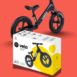 Black balance bike with 'velo' branding on a red background, next to its packaging box.